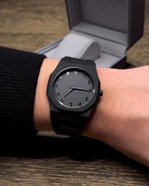 Minimalist black wristwatch with Arabic numerals on wrist, watch box in background